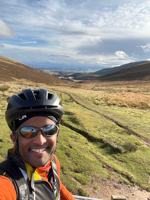 Ahmed, wearing black helmet and sports sunglasses, taking a selfie on his bike. Behind him is a bike path that leads to a reservoir surrounded by scenic views and hills.