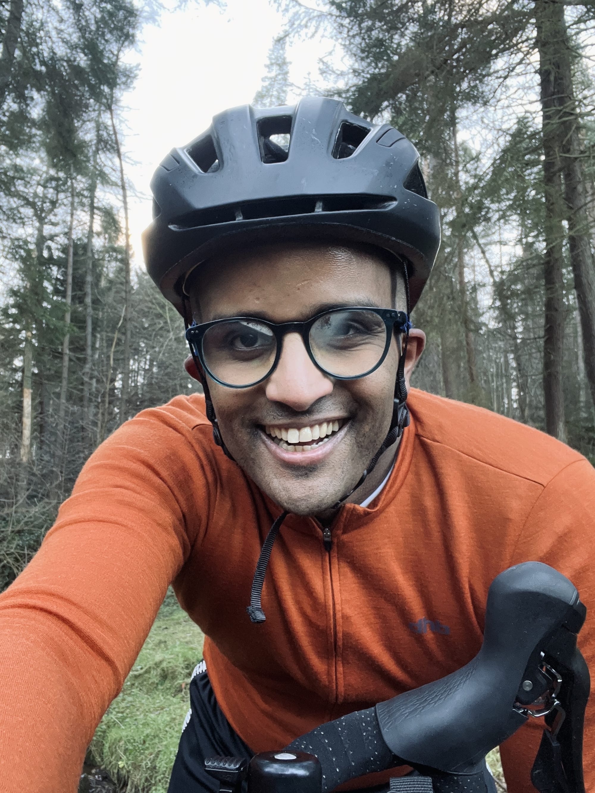 Ahmed, wearing an orange cycling sweater and helmet, is on his bike and smiling towards the camera with the woodland behind him.