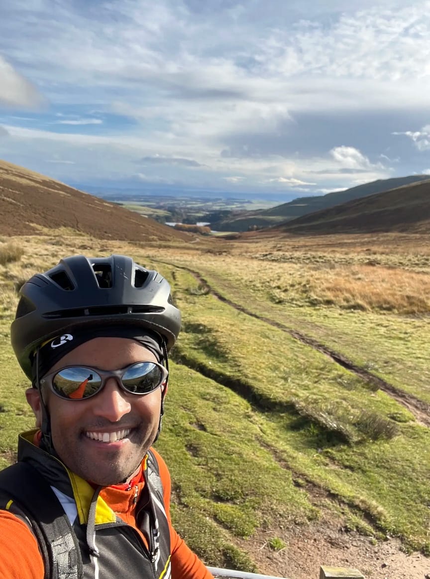 Ahmed on is bike, wearing sunglasses, helmet and cycling gears while taking a selfie on top of a hill. The backdrop is a beautiful view of the green Pentland moor with a reservoir in between two peaks.