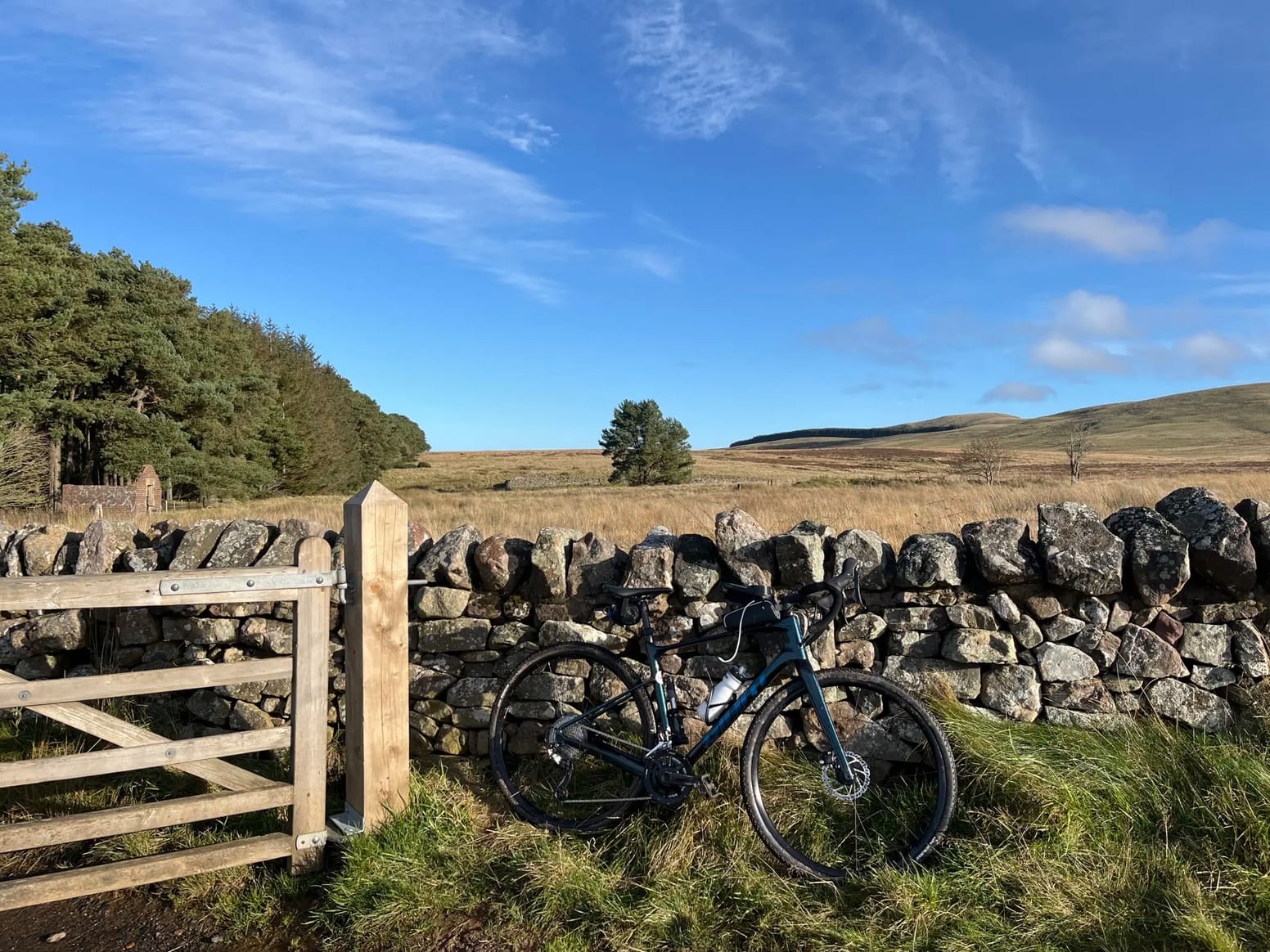 Bike resting against a stone wall beside a wooden fence with the backdrop of a yellow field with a tree in the middle, under a blue sky with specks of clouds.