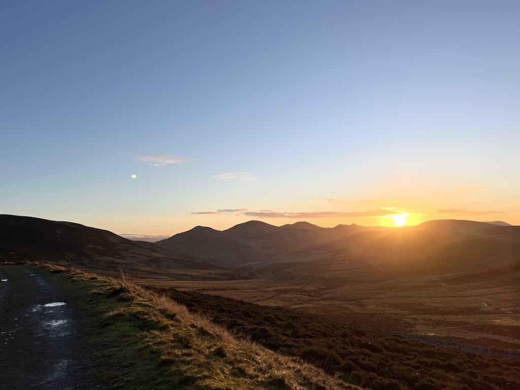 A view of the Pentland beside a wet gravel path. The sunset is slightly above the hills as it sets.