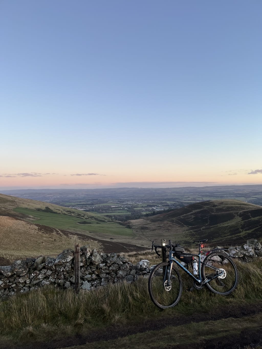 View of a valley beside the Pentland hills. The bike is leaning on a stone wall and standing beside a muddy path. The view goes out for miles with the sky slowly getting darker.